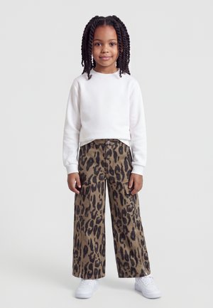 Jeune fille aux cheveux tressés portant une chemise blanche à manches longues, un pantalon large imprimé léopard et des baskets blanches, debout devant un fond uni.