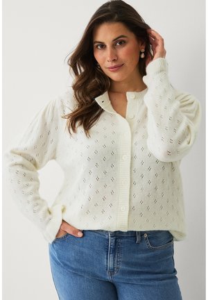 Kvinde iført hvid knapfast strikke-cardigan med små huller og blå jeans, poserer med den ene hånd i lommen og den anden, der rører ved håret.