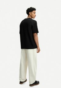 Črna teksturirana oversized majica, uparjena z belimi širokimi hlačami in črnimi čevlji, videna s hrbtne strani. Preprost dizajn in nevtralne barve.