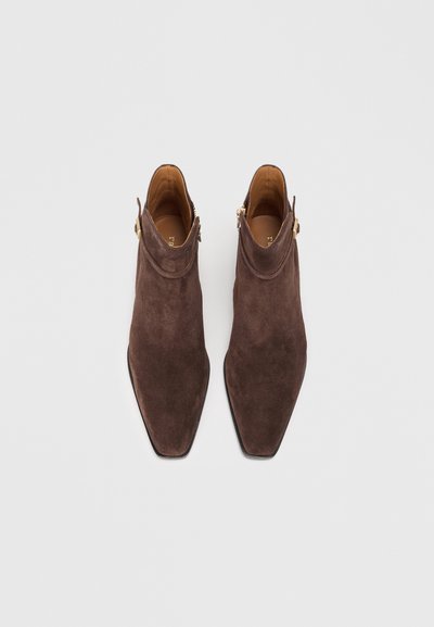Bottines en daim marron avec un design à bout pointu, présentant une discrète fermeture éclair sur le côté et des accents en métal doré. Semelle plate.
