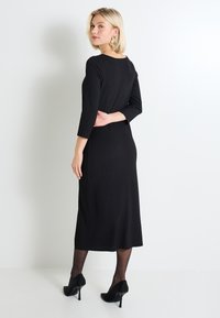 Vestido negro de manga tres cuartos, con silueta ajustada, escote redondo y dobladillo a la altura de la rodilla, combinado con tacones altos negros.