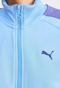 Giacca a zip di colore azzurro chiaro con collo a costine e dettagli viola sulle spalle. Presenta un logo viola ricamato sul lato sinistro del petto. Tessuto testurizzato.