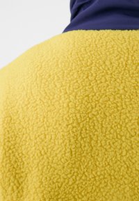 Fleecejack met een gele, textuur oppervlakte en een gladde marineblauwe bovenkant, die contrasterende kleuren en materialen laat zien.
