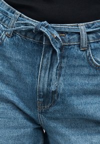 Μπλε τζιν παντελόνι με δέσιμο από ύφασμα σε φιόγκο στη μέση, ορατές μπροστινές τσέπες και μαύρο τοπ μαζεμένο μέσα.