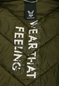 Innenansicht einer kurzen, olivgrünen, gepolsterten Jacke mit einem gesteppte Muster. Auf der Innenausstattung prangt großer, weißer Text: "TRAGE DAS GEFÜHL".