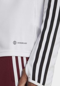 Maglia atletica bianca con strisce nere lungo le maniche, con logo "AEROREADY" vicino all'orlo. Tessuto morbido e texturizzato.