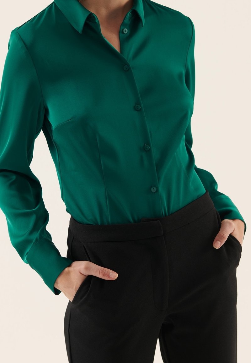 Femme portant un chemisier à manches longues vert émeraude boutonné, rentré dans un pantalon noir taille haute, mains dans les poches.
