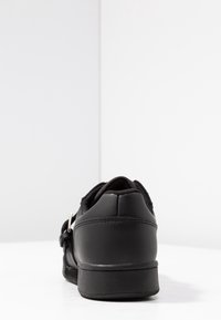 Chaussure noire avec un extérieur en cuir lisse, design arrondi, sangle réglable avec une quincaillerie en argent et semelle texturée.