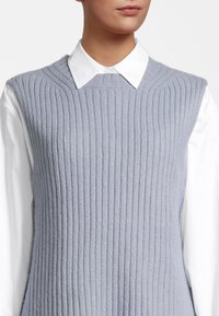 Un haut sans manches tricoté gris clair avec des côtes verticales, porté par-dessus une chemise blanche à col; texture douce et encolure ras du cou.