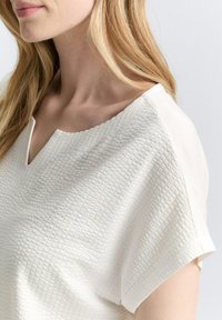 Blouse blanche en tissu texturé et à motifs. Présente un décolleté en V et des manches courtes avec des accents lisses.