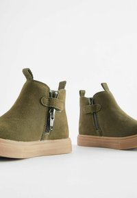 Botine din suede verde măsliniu, cu închidere prin fermoar, cu mânere din material textil și tălpi din cauciuc bej, având o textură netedă și un design minimalist.