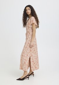 Woman with long curly hair wearing a beige floral midi dress with short sleeves and side slit, paired with black low-heeled slingback shoes.