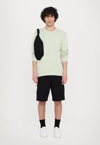 Felpa verde menta, pantaloni corti neri e sneakers bianchi. Borsa a tracolla nera con una cinghia. Modello in piedi contro uno sfondo bianco semplice.