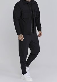 Svart zip-up jacka med två framfickor, kombinerad med matchande byxor och vita sneakers. Slät tyg med en figursydd design.