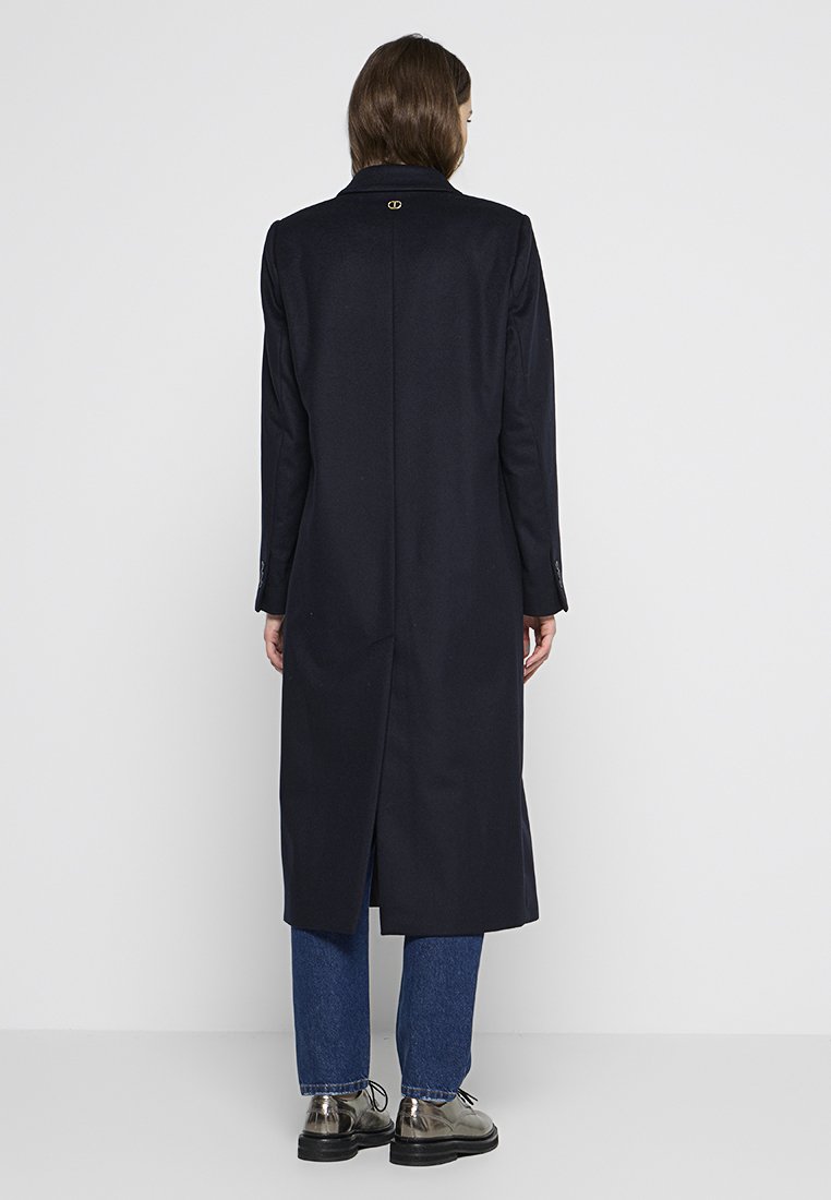 Marineblauwe jas met een gespleten achterkant, op maat gemaakt ontwerp, lange mouwen, enkel knopenmanchetten en een logo-accent bij de hals; gedragen over blauwe spijkerbroeken en schoenen.