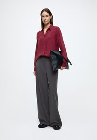 Dieprode blouse met knopen en een gestructureerd patroon, gecombineerd met grijze wijde broekspijpen en een zwarte draagtas. Glanzende zwarte schoenen maken de look compleet.