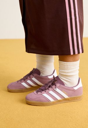 Pieds portant des baskets Adidas Gazelle roses avec des rayures blanches, des chaussettes blanches et un short marron avec des rayures roses, posés sur un sol jaune.
