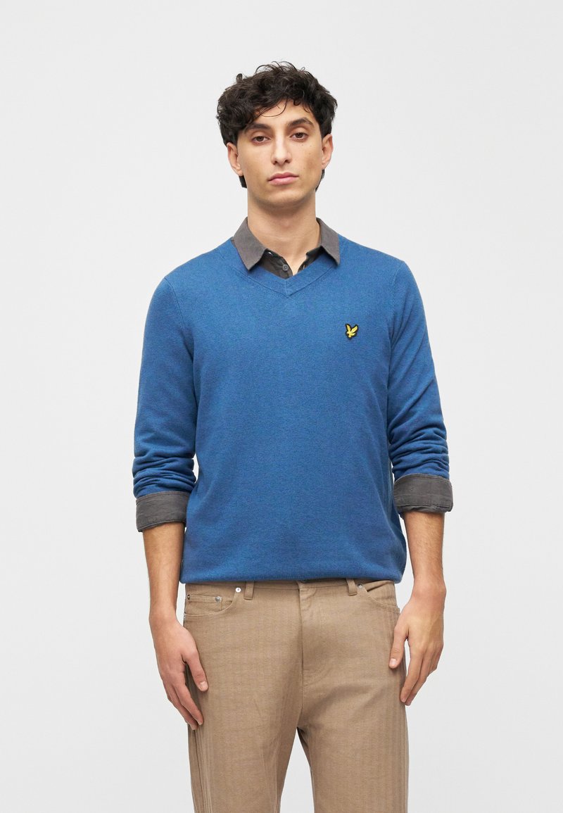 Kék V-nyakú pulóver kötött anyagból, szürke ing fölött, gallérral. Kis sárga logó található a mellkason, az ujjak felhajtva.