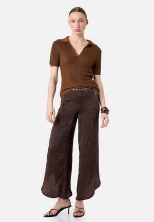 Mujer con un top de punto marrón de manga corta, pantalones anchos de pierna ancha texturizados oscuros con un cinturón de cadena dorada y sandalias de tacón negras con puntera abierta.