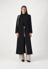 Cappotto nero con silhouette lunga e dritta, tasche laterali e design doppiopetto. Il look è completato da un top nero aderente e pantaloni beige a gamba larga.