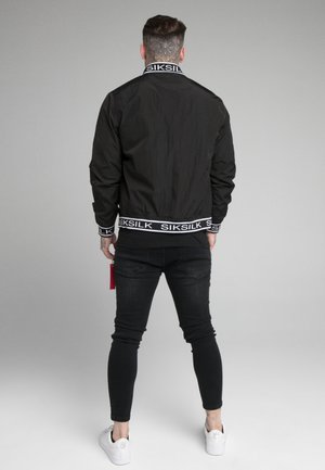 Uomo con un tatuaggio floreale sul collo, che indossa una giacca nera SikSilk, jeans skinny neri e sneakers bianche, in piedi contro un sfondo neutro.