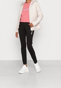 Vaalean beige puffatakki hupulla, vaaleanpunainen collegepaita kirjoituksella, mustat slim-fit-housut ja valkoiset tennarit neutraalilla taustalla.