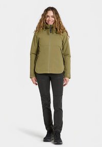 Olijfgroene softshell jas met ritssluiting aan de voorkant, zijzakken en een hoge kraag, gecombineerd met zwarte stretchbroek en robuuste zwarte laarzen.