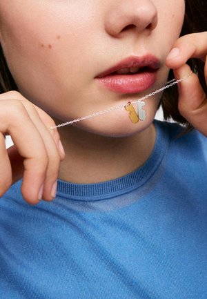 Joven con piel clara y labios ligeramente separados sostiene un collar de plata con piezas de puzzle entrelazadas de oro y plata.
