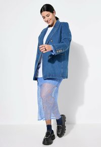 Blazer de denim azul con botones de doble botonadura, superpuesto a una falda ligera de encaje azul claro. Zapatos de plataforma negros gruesos y calcetines de rayas.