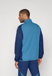 Blaues und navyfarbiges Sportjacke mit hohem Kragen, elastischen Bündchen und Reißverschlusstaschen; glatte Textur und kontrastierendes Paneeldesign an den Schultern.