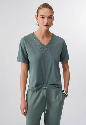 Ženska, oblečena v turkizno modro majico z V-izrezom in kratkimi rokavi, okrašeno z drobnimi kristali, ter ujemajoče, z vrvico na pasu pritrjene hlače, dopolnjena z velikimi srebrnimi uhani v obliki obročev in zapestnicami.