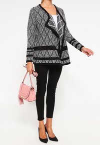 Svartvit mönstrad cardigan med draperad front, bärs över en vit topp, ihop med svarta byxor och en rosa handväska.