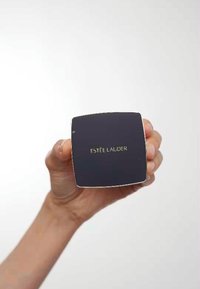 Firkantet pudder med en glossy mørkeblå finish, sølvdetaljer og gull "Estée Lauder" logo, holdt i en hånd, mot en lys bakgrunn.