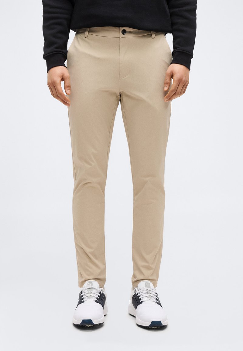 Homme portant un pantalon beige ajusté et des baskets blanches avec des accents noirs, debout devant un fond clair uni.