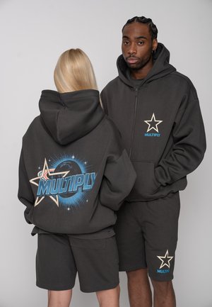 Zwei Models tragen schwarze Kapuzenpullover und Shorts mit dem Schriftzug „MULTIPLY“ und Sternzeichnungen; die Frau zeigt das Design auf dem Rücken, der Mann steht vorn mit den Händen in den Taschen.