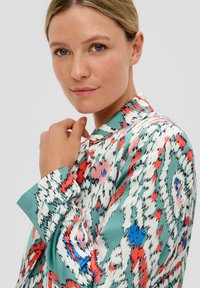 Seidenbluse mit einem lebhaften Muster aus Rot, Blau und Grün auf einem weißen Grund, mit einer weichen Textur und einem klassischen Kragen.