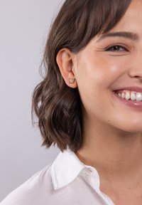 Goldene, schmetterlingsförmige Ohrringe mit kleinen Akzenten, getragen an dem Ohr eines Models. Das Model hat dunkles, welliges Haar und trägt ein weißes Hemd mit Kragen.