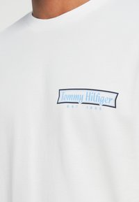 Hvit kortermet t-skjorte laget av bomull, med et blått og svart merkevarelogo på brystet og teksten "EST 1985" under.