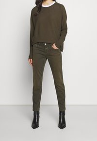 Haut à manches longues vert olive avec un col blanc et un pantalon slim assorti vert olive. Le modèle porte des bottines noires à bout pointu.