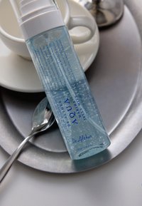 Bottiglia trasparente di Dr. Althea Aqua Marine Jelly Mist con bolle, posata su una tazza bianca con piattino su un vassoio grigio con un cucchiaio.