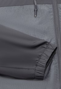 Grijze windbreaker jas met een gestructureerd bovenstuk, een glad onderstuk, elastische manchetten en een ritsluiting aan één zijde.