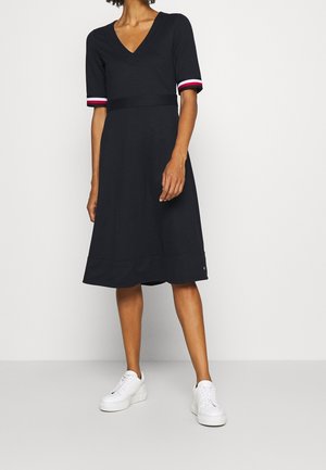 Femme portant une robe noire mi-longue à col en V avec des manches courtes bordées de rouge, blanc et bleu, assortie à des baskets blanches, debout sur un sol gris.