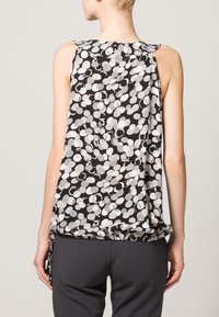 Beżowa bezrękawnik w czarno-białym materiale, z okrągłym wzorem i luźnym krojem. Okrągły dekolt z plisowaniem z tyłu.