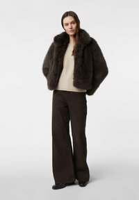 Brązowa kurtka z futerkiem syntetycznym o szerokim kołnierzu, beżowy sweter dzianinowy oraz brązowe spodnie o szerokich nogawkach. Czarne buty dopełniają stylizację na jednolitym tle.