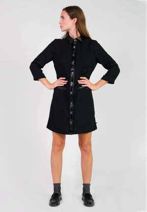 Schwarzes, strukturiertes Kleid mit Kragen, vorderen Knöpfen, zwei Taschen und Dreiviertelärmeln. Kombiniert mit grauen Socken und schwarzen Schuhen.