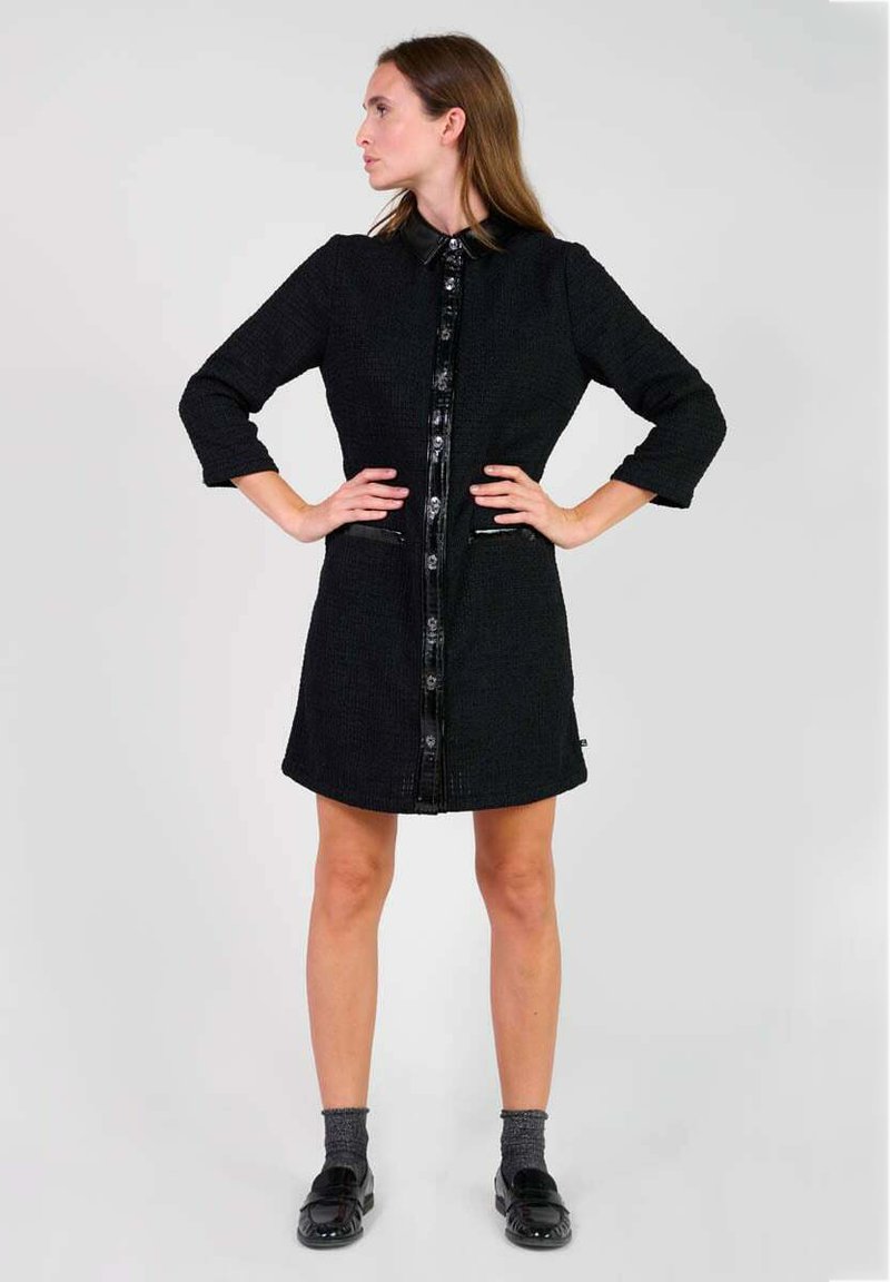 Vestido negro texturizado con cuello, botones en el frente, dos bolsillos y mangas de tres cuartos. Combinado con calcetines grises y zapatos negros.