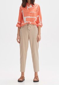 Chemise boutonnée corail avec un motif abstrait blanc, pantalon beige à cordon, et sandales marron avec une semelle épaisse. Style décontracté en tissu léger.