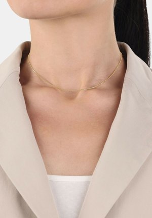 Collar de oro con un diseño de cadena delicada, llevado sobre un cuello desnudo contra una chaqueta de color claro y una blusa blanca.