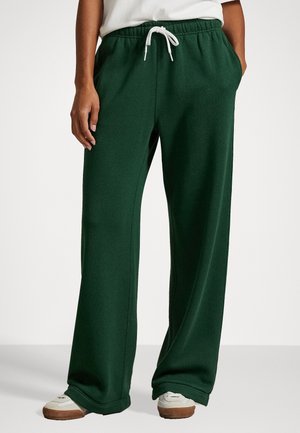 Person iført mørkegrønne vidde-sweatpants med hvid snøre og hvide sneakers, stående foran en ensfarvet baggrund.