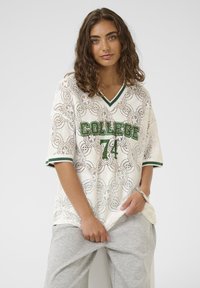 Mlada ženska s kodrastimi lasmi nosi belo čipkastem "College 74" sivo-zelenim trimom in svetlo sivo trenirko, pozira na enobarvni podlagi.